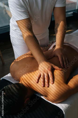 Vertical top view shot of professional male masseur with strong tattooed hands massaging back and shoulders of muscular sports man lying on stomach, at massage table on background of sunlight.