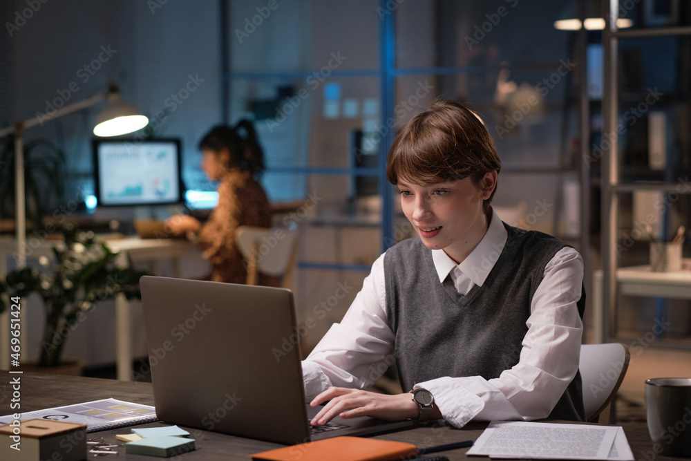 Young businesswoman sitting at the table typing on laptop and working online at office till late evening