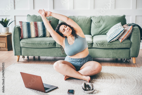 Curly haired overweight young woman in top and shorts turns on online yoga training and practices exercises on floor mat against green sofa by wall