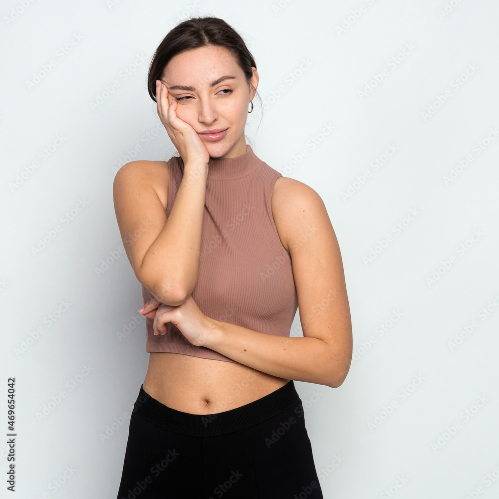 Obraz premium Cute young woman standing in short turtleneck, holding palm on cheek and leaning face on it, being bored and tired of uninteresting talks, feeling indifferent to what happening over gray wall