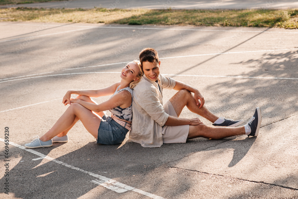 summer holidays and people concept - happy young couple sitting back to back on basketball playground