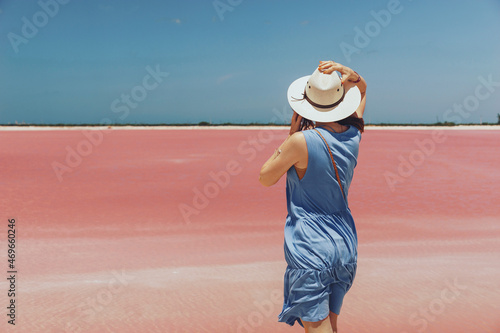 A young woman in a hat and blue dress stands against the backdrop of a salty pink lake in Mexico, seen from the back. Amazing view of Pink Lake, popular tourist spot in Mexico