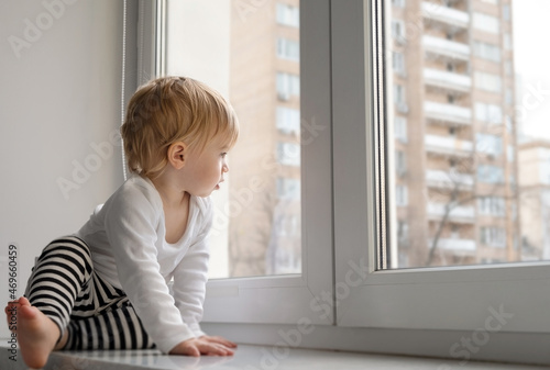 Cute caucasian blonde baby girl about 1,2 years old sitting on sill looking through window at street autumn view.Infant,toddler,kid safety,secuity.