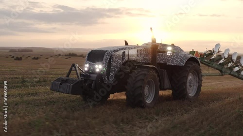 An unmanned tractor drives through a field and plows the land with a plow at sunset