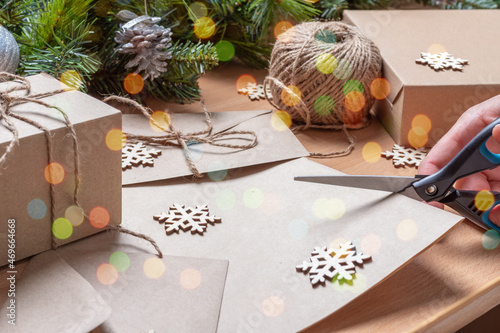 hand cutting recycled paper with scissors on the table with artificial fir tree with cones, envelope, twine, and wooden snowflakes