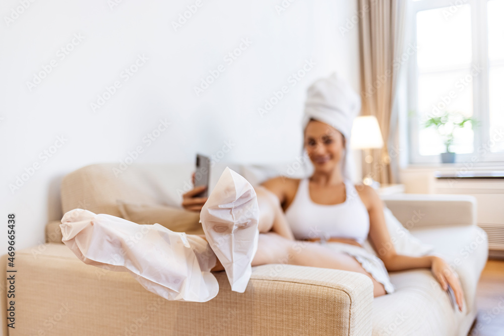 Close-up of woman's feet wrapped in moisturizing socks during home pedicure. Young happy woman texting on mobile phone while using moisturizing socks on her feet after having pedicure at home. Close-up of woman's feet wrapped in moisturizing socks during home pedicure. Young happy woman texting on mobile phone while using moisturizing socks on her feet after having pedicure at home.