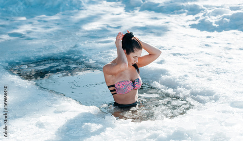 Girl with bikini and a watch in frozen lake ice hole. Woman hardening the body in cold water. Good immunity is protection against many diseases. Vintage color filter photo