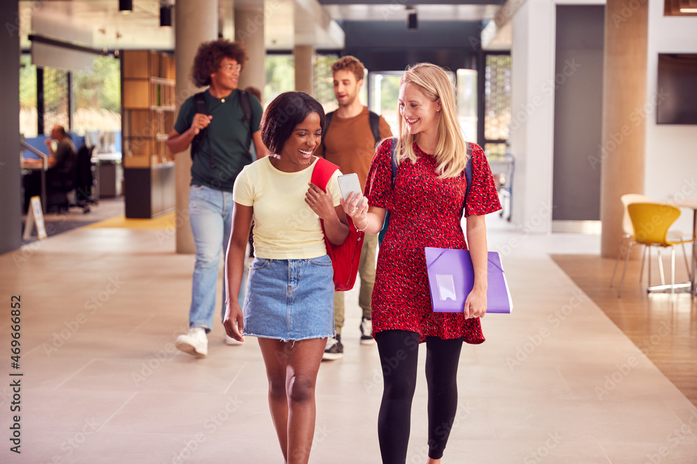 Two Female Students In Busy University Or College Building Looking At Mobile Phone Two Female Students In Busy University Or College Building Looking At Mobile Phone