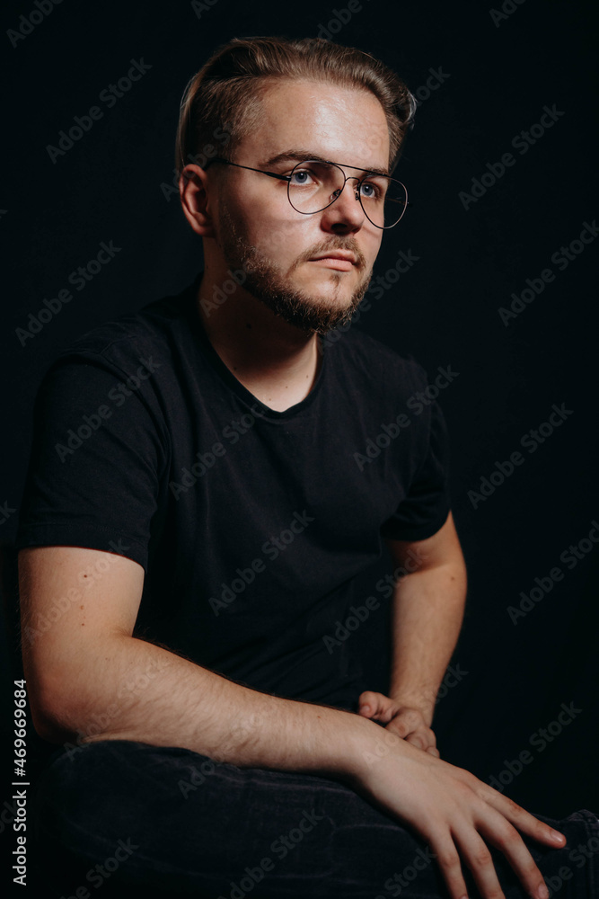 A important guy in a T-shirt and glasses and with a modern haircut and beard sits in the studio on a black background and with a serious inquisitive look A important guy in a T-shirt and glasses and with a modern haircut and beard sits in the studio on a black background and with a serious inquisitive look