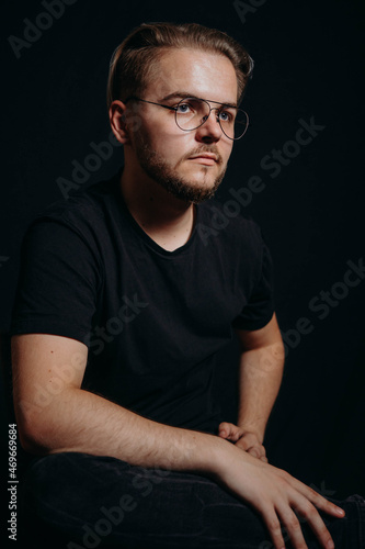 A important guy in a T-shirt and glasses and with a modern haircut and beard sits in the studio on a black background and with a serious inquisitive look 