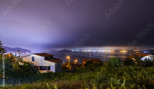 Ría de Pontevedra de noche con niebla y ciudad de fondo con luces y bateas de mejillones. Casa entre la vegetación y isla al fondo