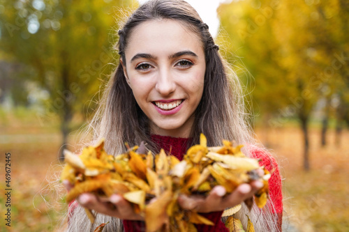 Happy smiling girl holding yellow leaves in hands. Portrait of young woman on background of park. Autumn concept