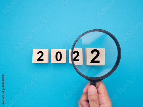 Hand of holding a magnifying glass looking to ideas for the 2022 year. Close-up photo. Top view of wooden blocks with words 2022. Space for text