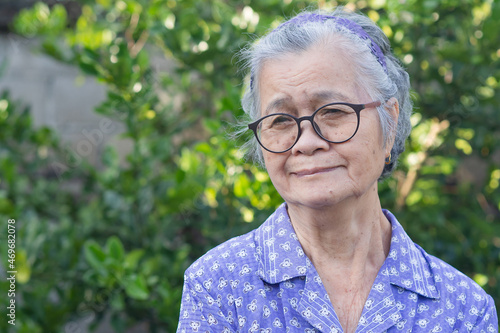 Portrait of an elderly Asian woman with short gray hair, wearing glasses, and looking away with a smile while standing outdoor. Aged people and relaxation concept