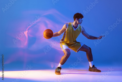 Dynamic portrait of professional basketball player training with ball isolated on blue studio background in mixed neon light.