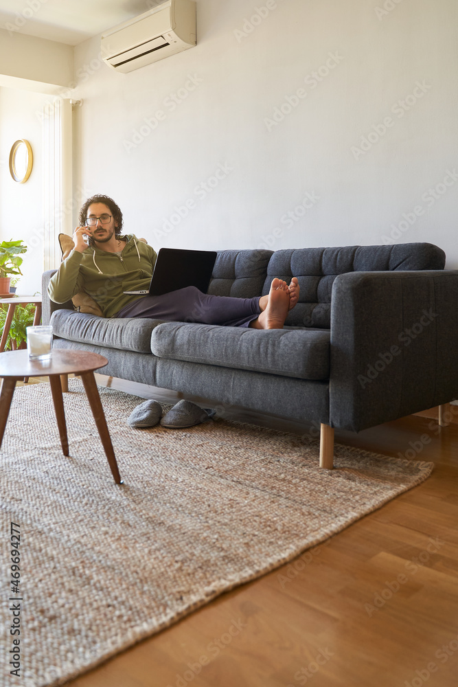 Vertical shot of young man speaking on the phone while laying on the sofa and using laptop Vertical shot of young man speaking on the phone while laying on the sofa and using laptop