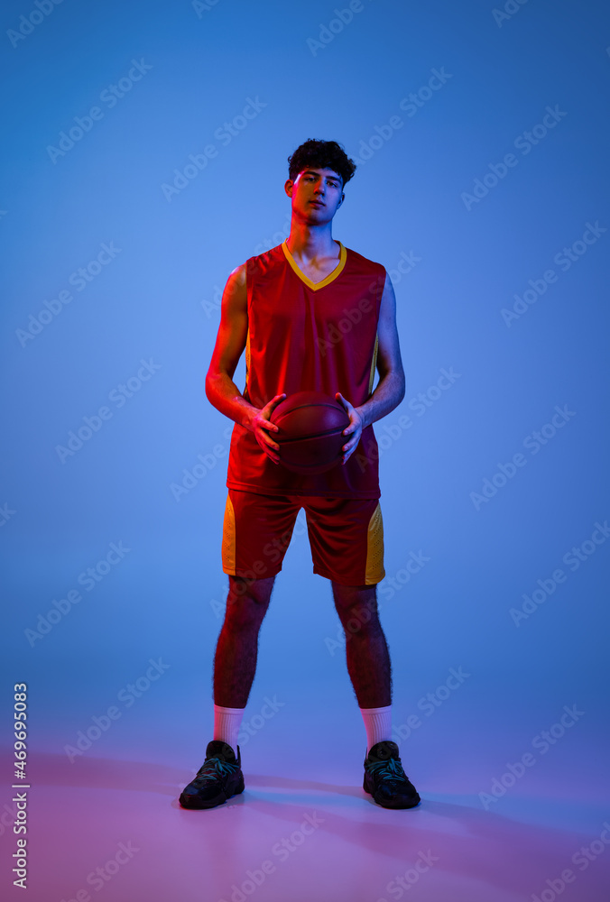Portrait of professional basketball player posing with ball isolated on blue studio background in neon light.