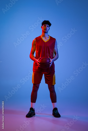 Portrait of professional basketball player posing with ball isolated on blue studio background in neon light.