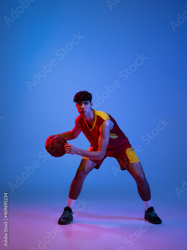 Studio shot of young man, professional basketball player playing basketball isolated on blue background in neon light. © master1305