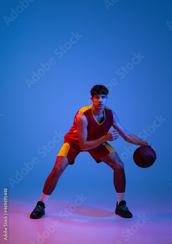 Studio shot of young man, professional basketball player playing basketball isolated on blue background in neon light.