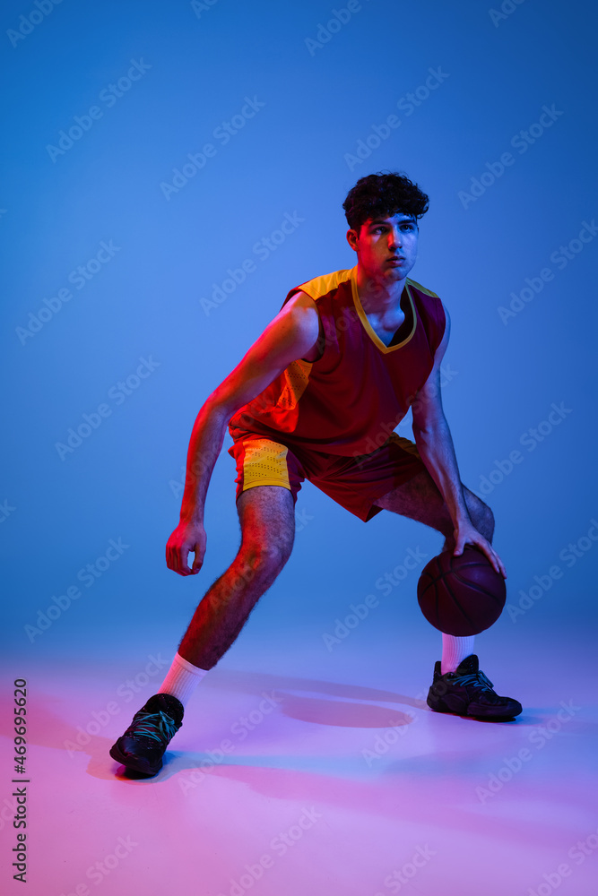 Studio shot of young man, professional basketball player playing basketball isolated on blue background in neon light.