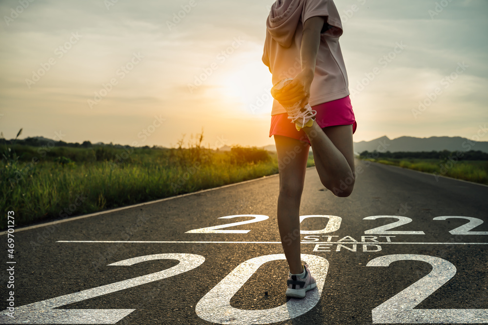 New year 2022 or start straight concept.word 2022 written on the asphalt road and athlete woman runner stretching leg preparing for new year at sunset.Concept of challenge or career path and change.
 - obrazy, fototapety, plakaty 