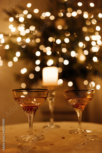 Two glass of sparkling wine and a candle stand on the table in front of the Christmas tree and blurred lights in the evening