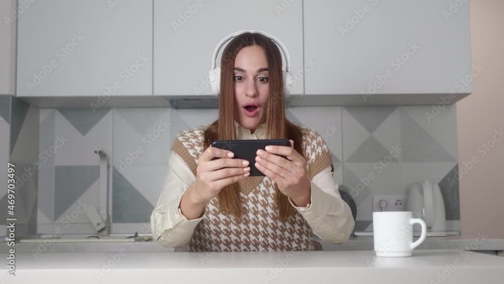 Amazed woman playing smartphone shocked, saying WOW. Female in headphone surprised to camera over kitchen background. Amazed woman playing smartphone shocked, saying WOW. Female in headphone surprised to camera over kitchen background.