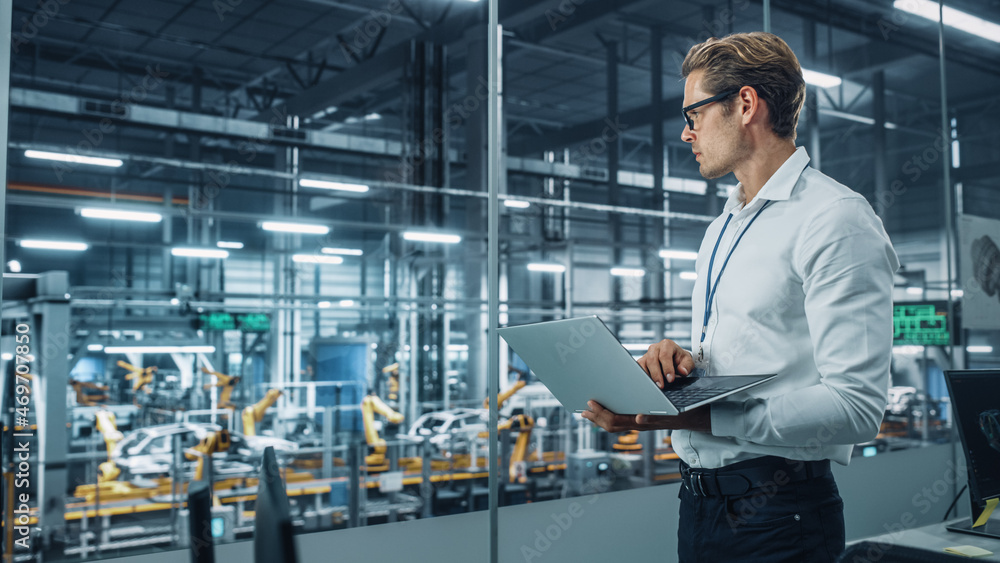 Handsome Engineer in Glasses and White Shirt Using Laptop Computer and Looking Out of the Office at a Car Assembly Plant. Industrial Specialist Working on Vehicle Design in Technological Facility. Handsome Engineer in Glasses and White Shirt Using Laptop Computer and Looking Out of the Office at a Car Assembly Plant. Industrial Specialist Working on Vehicle Design in Technological Facility.