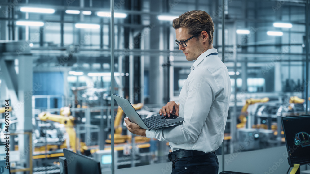 Handsome Engineer in Glasses and White Shirt Using Laptop Computer and Looking Out of the Office at a Car Assembly Plant. Industrial Specialist Working on Vehicle Design in Technological Facility. Handsome Engineer in Glasses and White Shirt Using Laptop Computer and Looking Out of the Office at a Car Assembly Plant. Industrial Specialist Working on Vehicle Design in Technological Facility.
