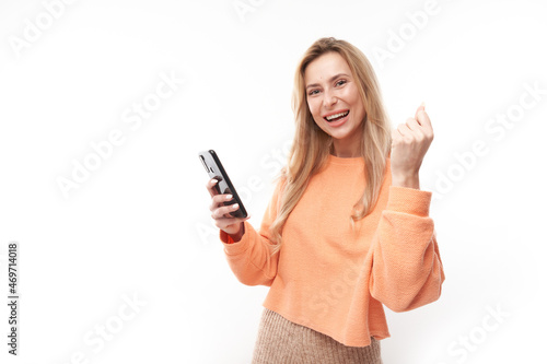 lucky smiling blond girl holding smartphone in casual isolated in white studio background received joyful message, celebrating victory