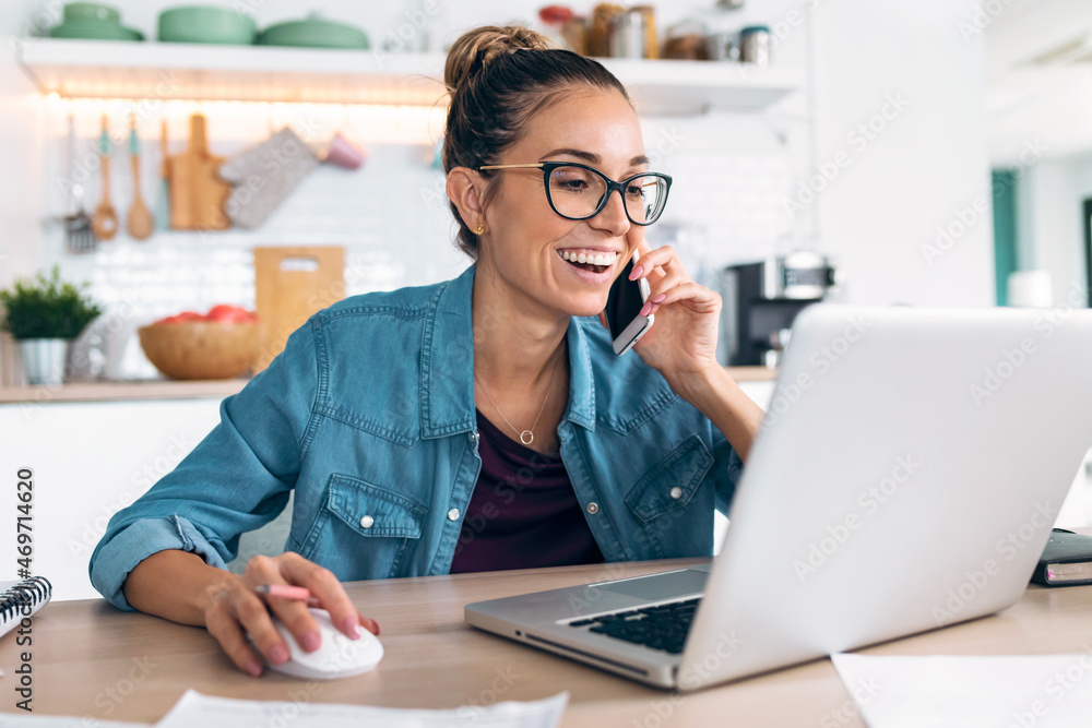 Smiling business woman working with laptop while talking with smartphone in the kitchen at home. Smiling business woman working with laptop while talking with smartphone in the kitchen at home.