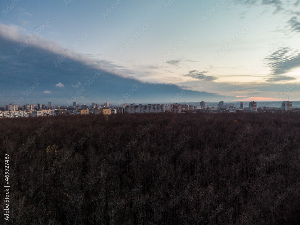 Aerial sunset evening city above autumn forest near residential Pavlove Pole district in Kharkiv city. Gray multistory buildings with scenic blue cloudy sky on horizon