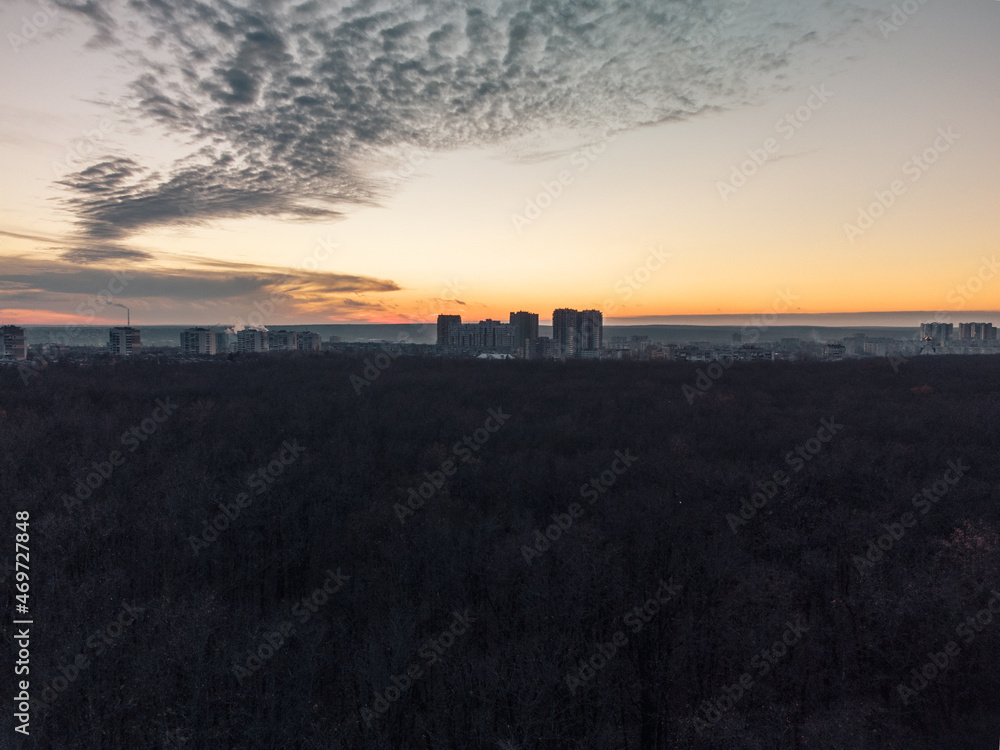 Aerial sunset evening view above dark autumn forest near residential Pavlove Pole district in Kharkiv city. Gray multistory buildings with scenic orange cloudy sky on horizon
