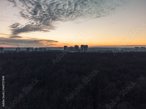 Aerial sunset evening view above dark autumn forest near residential Pavlove Pole district in Kharkiv city. Gray multistory buildings with scenic orange cloudy sky on horizon