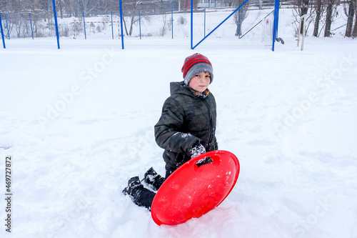 little boy playing in the snow, wearing a red hat, snowy, with a plasma plate for a slide,s kating rink  new year, christmas photo