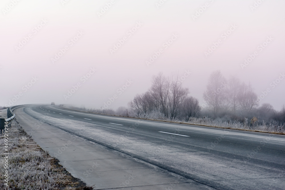 A road with a sharp bend in the early morning in frosty foggy weather. Grass, trees in frost and fog along the road. A road with a sharp bend in the early morning in frosty foggy weather. Grass, trees in frost and fog along the road.