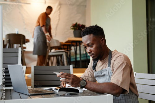 Young man wearing apron sitting at table in front of laptop in modern cafe taking online orders from customers for delivery