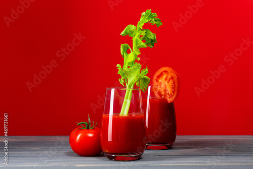 two glasses of tomato juice with celery and a whole tomato stand on a table made of gray boards on a red background