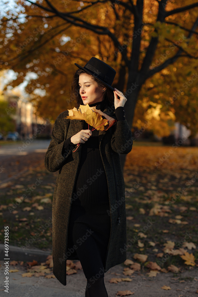 Young beautiful pregnant woman in a hat with dark hair in a black tight dress and coat posing on an autumn meadow in the park Young beautiful pregnant woman in a hat with dark hair in a black tight dress and coat posing on an autumn meadow in the park