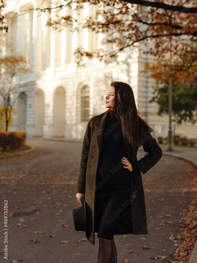 Young beautiful pregnant woman with dark hair in a black tight dress and coat posing on an autumn meadow in the park