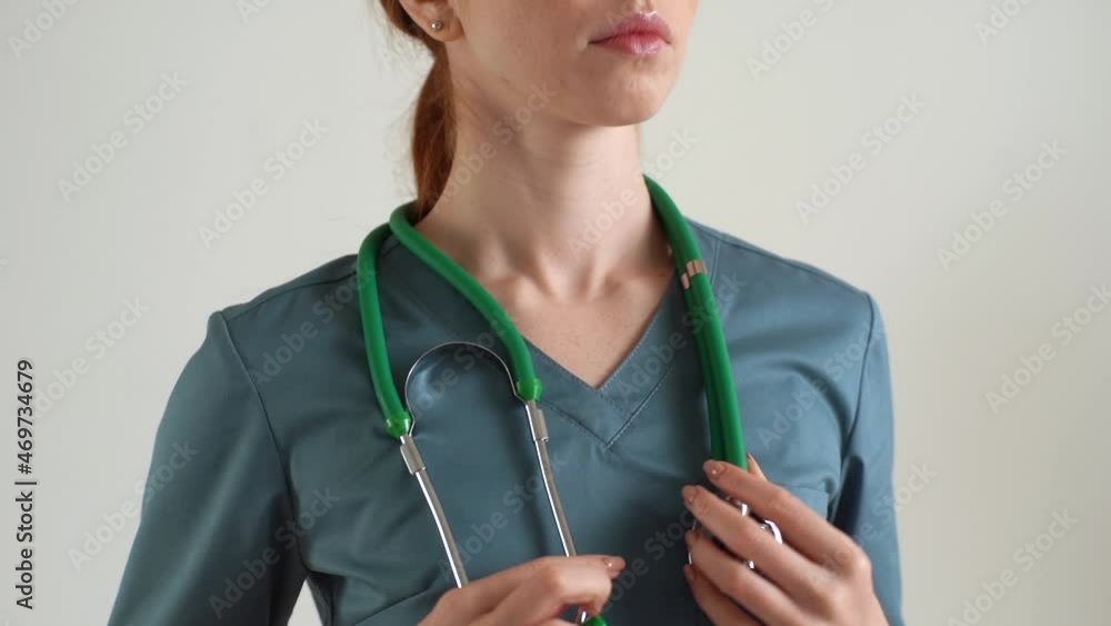 Close-up cropped shot of unrecognizable female practitioner in green medical uniform putting stethoscope around neck standing on white isolated background in studio. Shooting in slow motion.