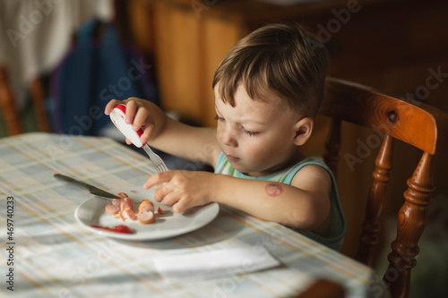 Boy sittyng by table and eating hot-dogs with fork