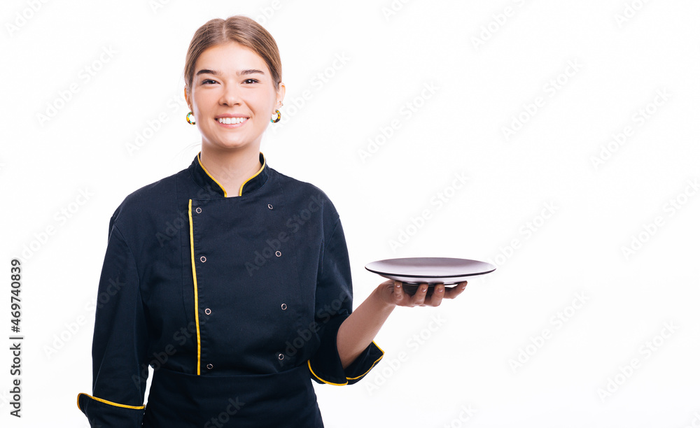 Joyful young woman chef standing over white background and holding empty black plate. Joyful young woman chef standing over white background and holding empty black plate.