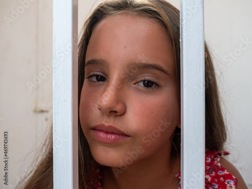 A cute blond Caucasian teenage girl with long hair and red dress sitting on the floor behind bars and looking at the camera