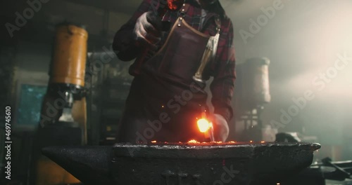 A strong man hits a metal workpiece that lies on an anvil. He holds her back with tongs. Sparks fly in different directions from the impact. The blacksmith is wearing a protective suit