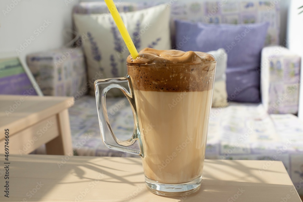Close up delicious milk frappe drink with dark coffee cream in transparent glass on light beige wooden table. Blurred background of cozy sofa with pillow cushions. Relaxation, holidays in sunny mood. Close up delicious milk frappe drink with dark coffee cream in transparent glass on light beige wooden table. Blurred background of cozy sofa with pillow cushions. Relaxation, holidays in sunny mood.