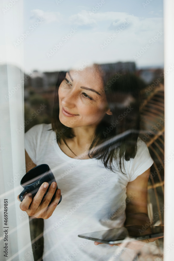 Smiled and positive woman looks out the window of her apartment or office. She is holding cup of coffee and tablet. Quarantine and social distancing during Covid-19 pandemic concept. Stay positive.