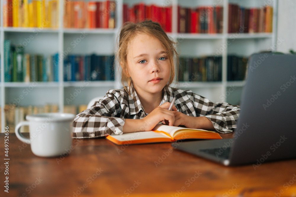 Close-up portrait of adorable pretty primary child school girl doing homework sitting at table with laptop and paper notebook, looking at camera. Cute pupil schoolgirl e-learning online using computer Close-up portrait of adorable pretty primary child school girl doing homework sitting at table with laptop and paper notebook, looking at camera. Cute pupil schoolgirl e-learning online using computer