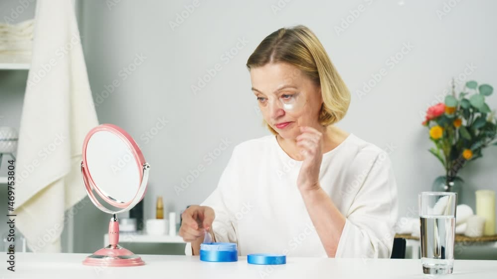 Old woman using eyes patches close-up, looking in mirror. Face skin care, beauty procedure, morning routine in bathroom. Cosmetology concept, cosmetic products. Old woman using eyes patches close-up, looking in mirror. Face skin care, beauty procedure, morning routine in bathroom. Cosmetology concept, cosmetic products.